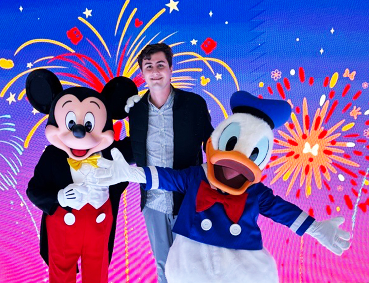 El pasante de The Walt Disney Company LATAM México, Guillermo, con blazer, camisa de cuello y jeans, sonríe junto a Mickey Mouse y el Pato Donald.